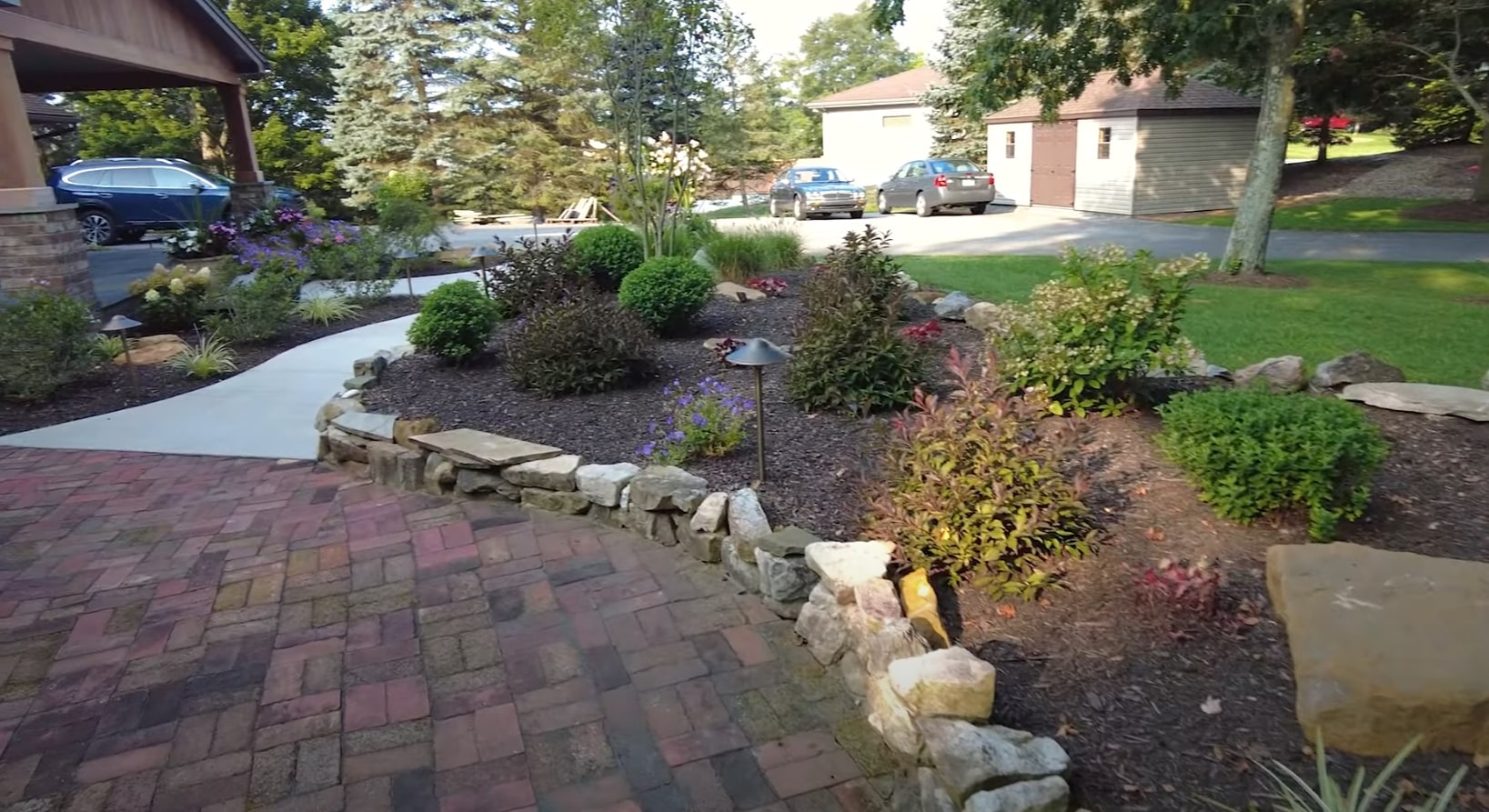 Complete Landscape Renovation - Stone Walls and Seasonal Plant Installation Front yard landscape renovation featuring natural stone retaining walls, colorful plantings, and brick paver driveway